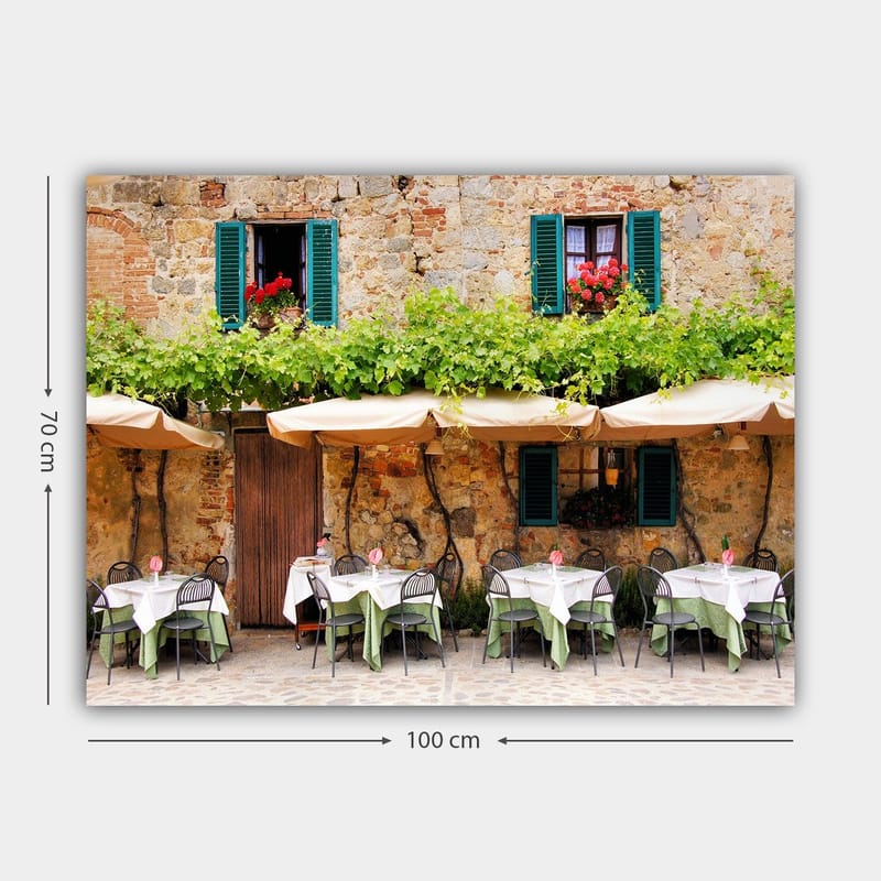 Canvastavla 70x100 cm - Charmig uteservering med vita bord och gröna dukar framför en gammal stenbyggnad med fönsterluckor i mörkgrön färg - Mörkgrön / Vit / Tegelröd - Inredning & dekor - Tavlor & konst - Canvastavla