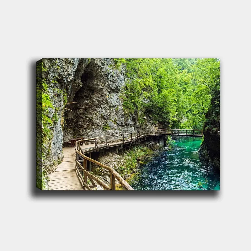 Canvastavla 70x100 cm - Naturskön vy av en träbro som slingrar sig genom en grönskande ravin vid en klar, turkos flod, Grön / Turkos / Grå