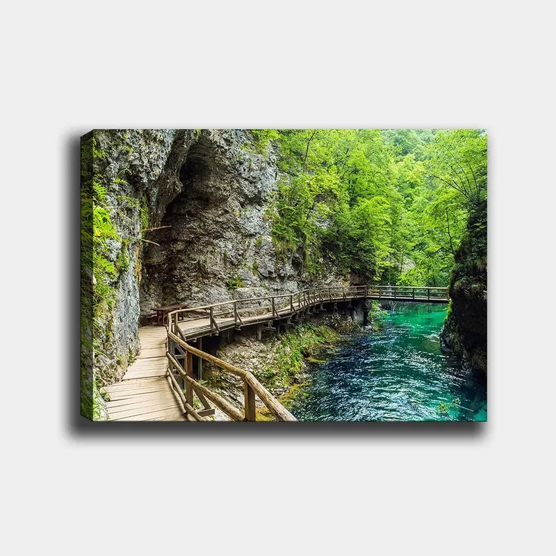 Canvastavla 70x100 cm - Naturskön vy av en träbro som slingrar sig genom en grönskande ravin vid en klar, turkos flod, Grön / Turkos / Grå